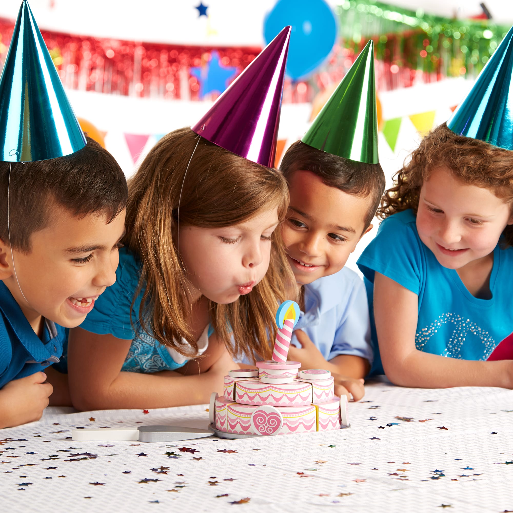 Children wearing colorful party hats at a birthday party, looking at a small cake with candles.
