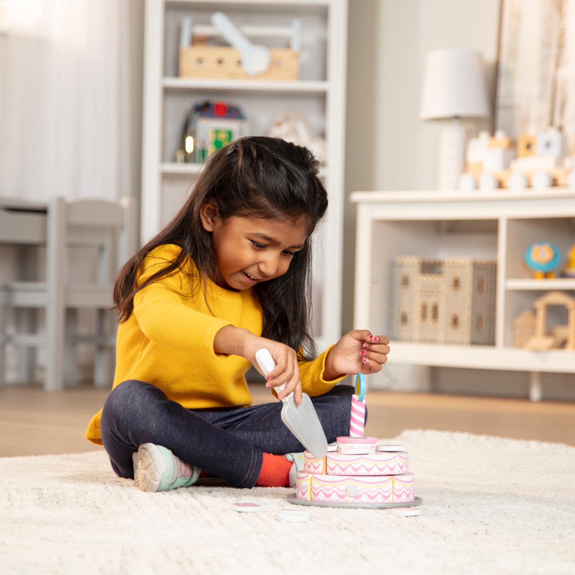 Young girl in a yellow sweater playing with a toy kitchen set in a room with shelves and toys.