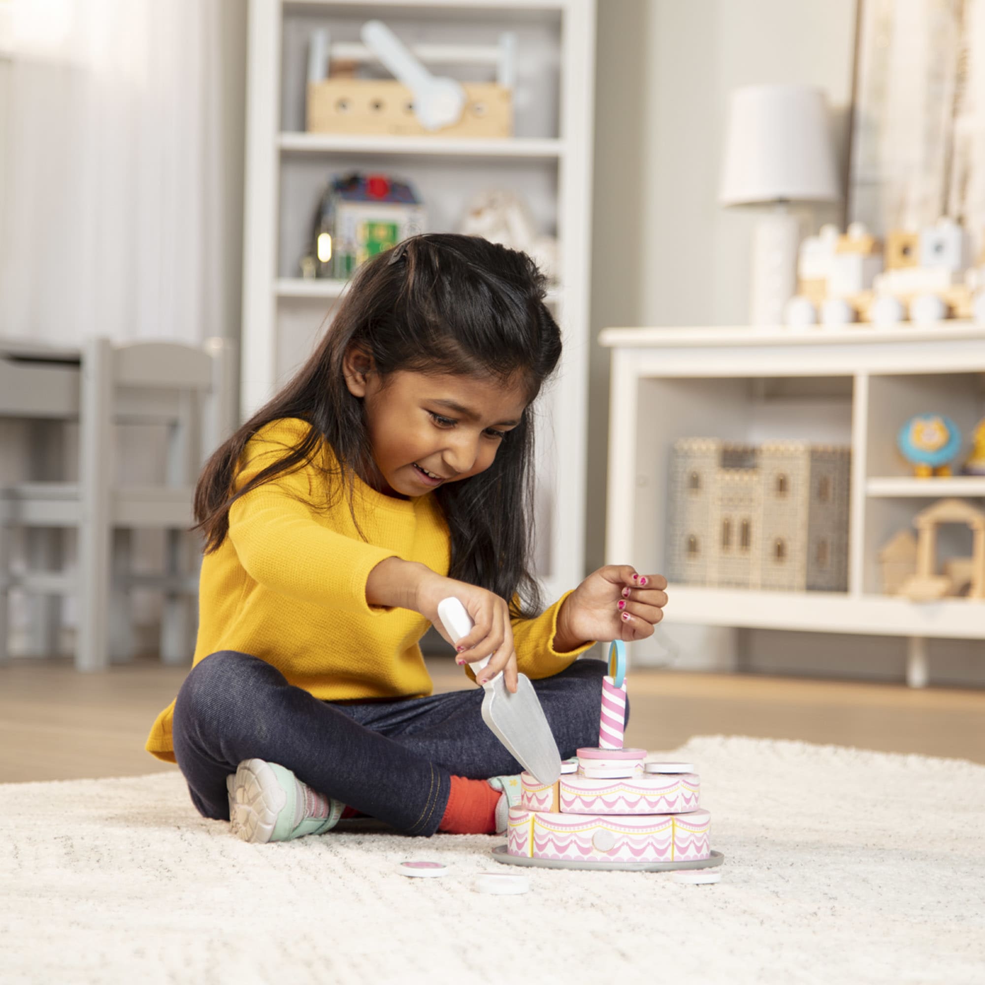 Young girl in a yellow sweater playing with a toy kitchen set in a room with shelves and toys.