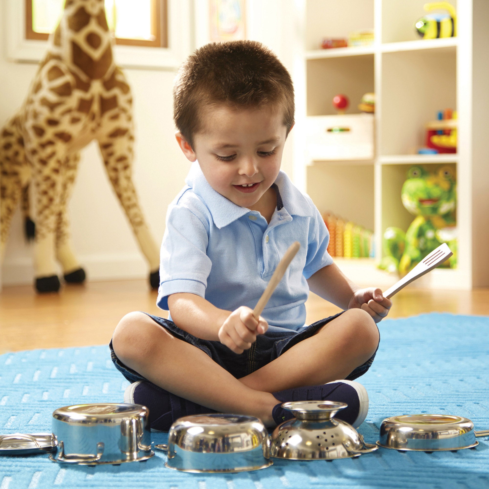 Child playing with pots and pans on a blue blanket in a room with toys and a giraffe.