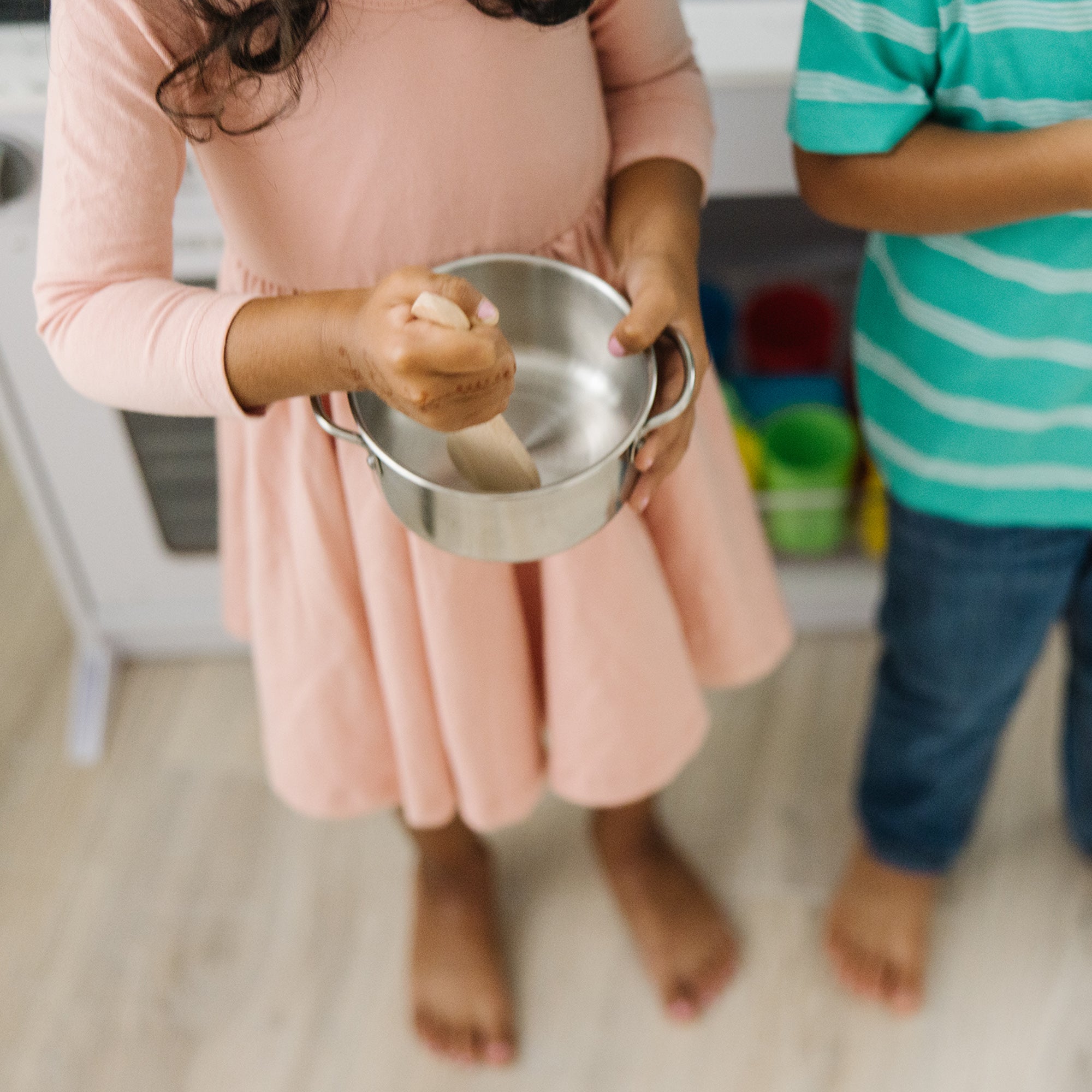 Child in a pink dress holding a metal bowl with a child in a striped shirt in the background.