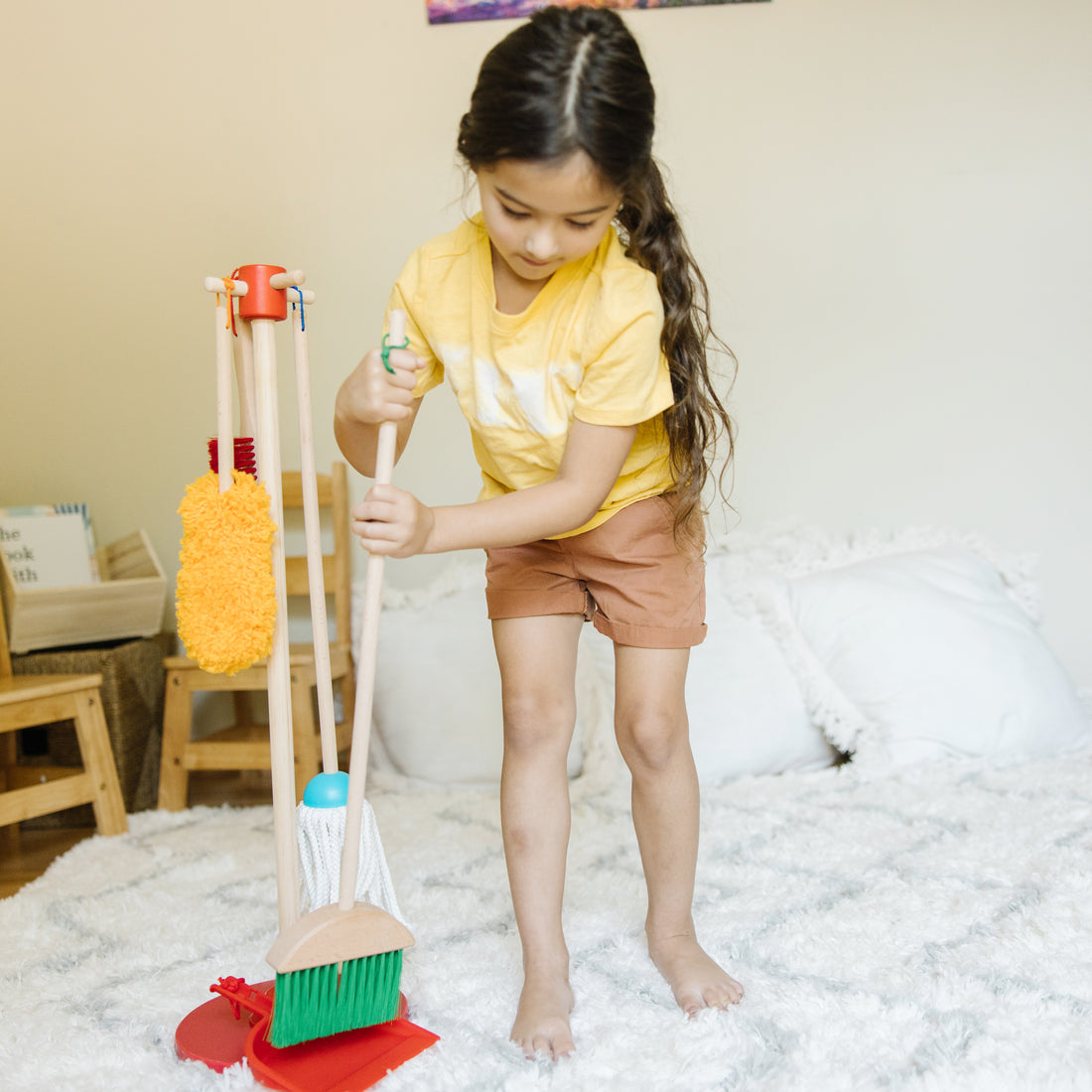 Child in a yellow shirt and brown shorts holding cleaning tools on a bed.