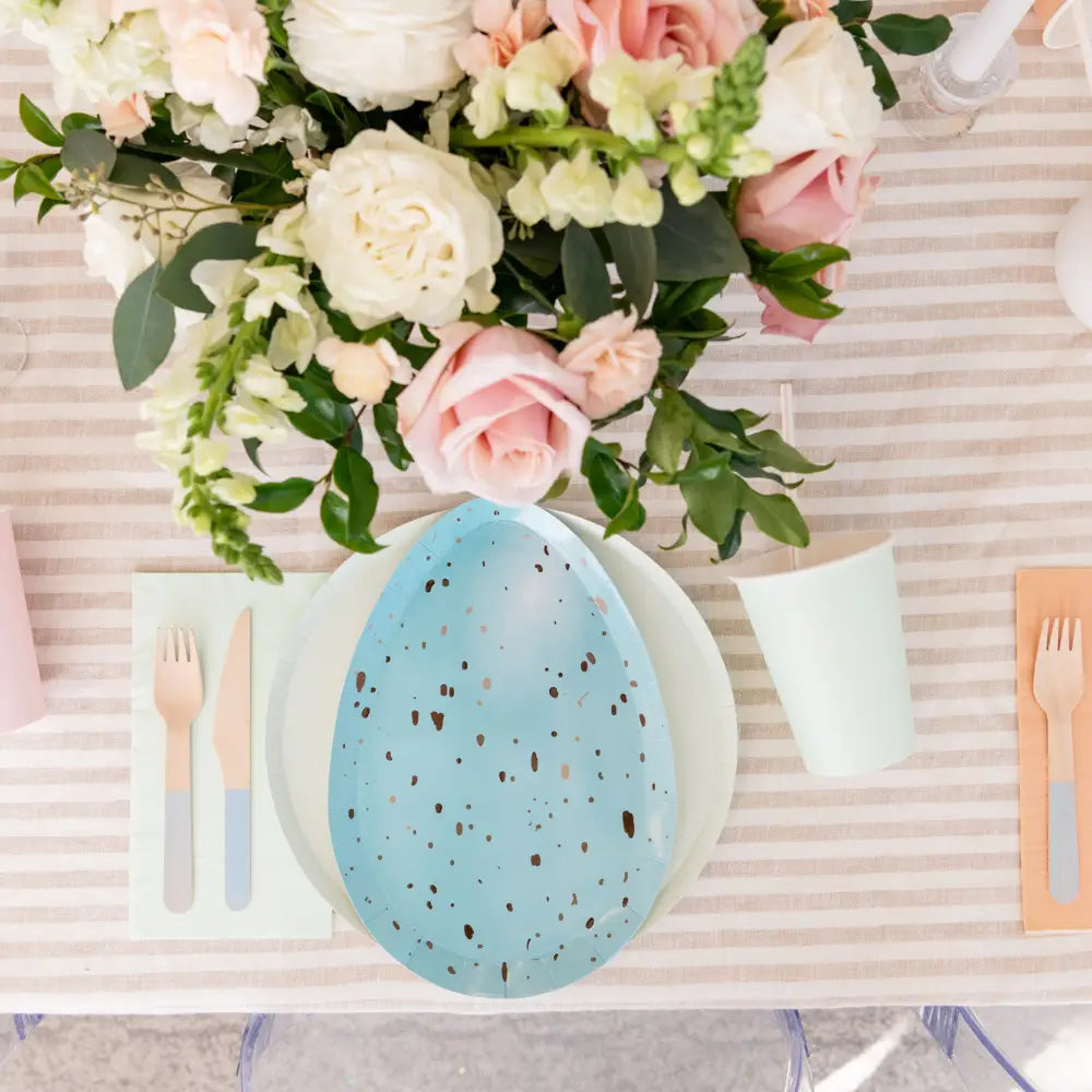 Table setting with a blue plate, pink and white flowers, and cutlery on a striped tablecloth.