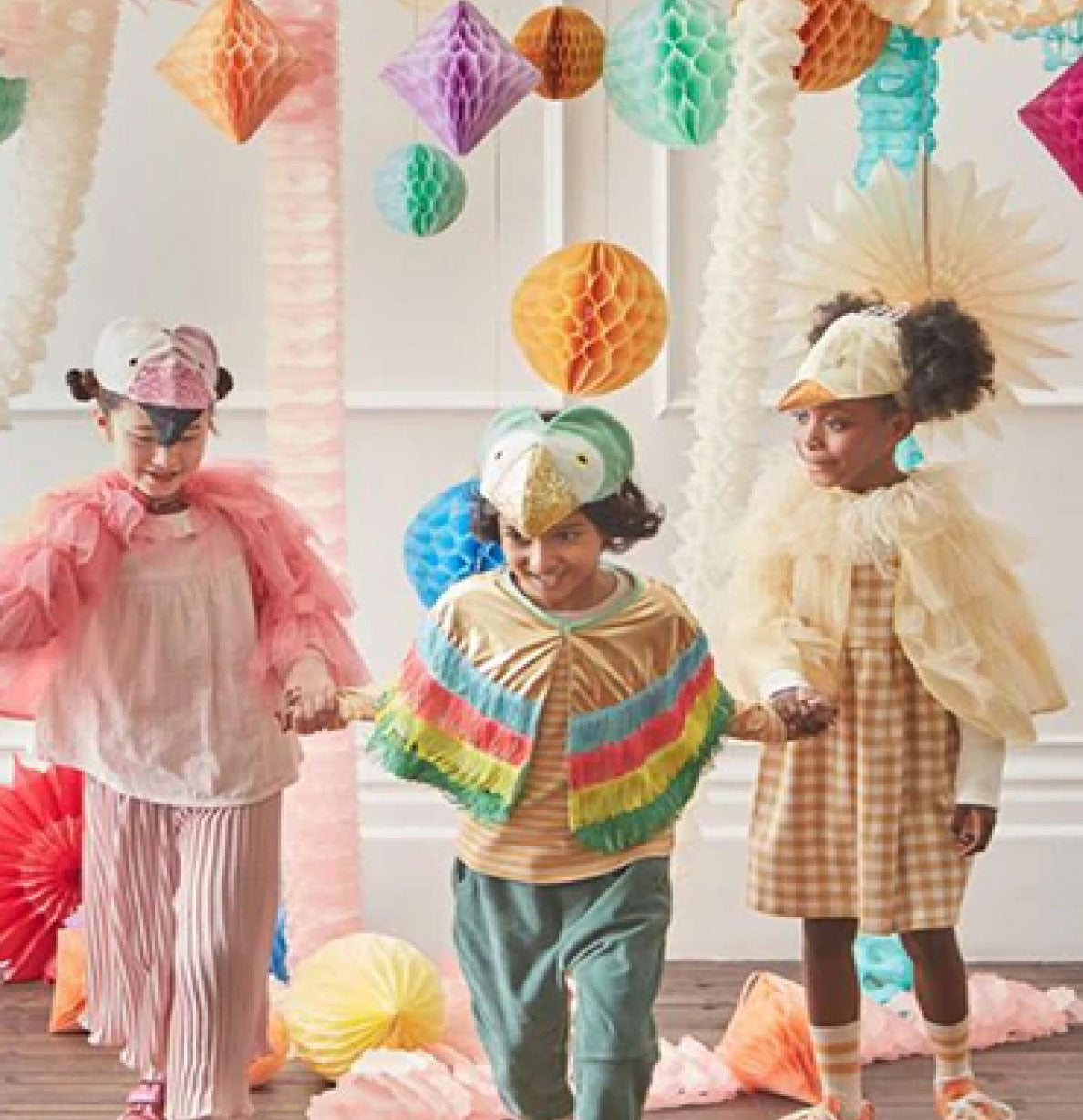 Three children in colorful outfits standing in a room with decorative paper lanterns.